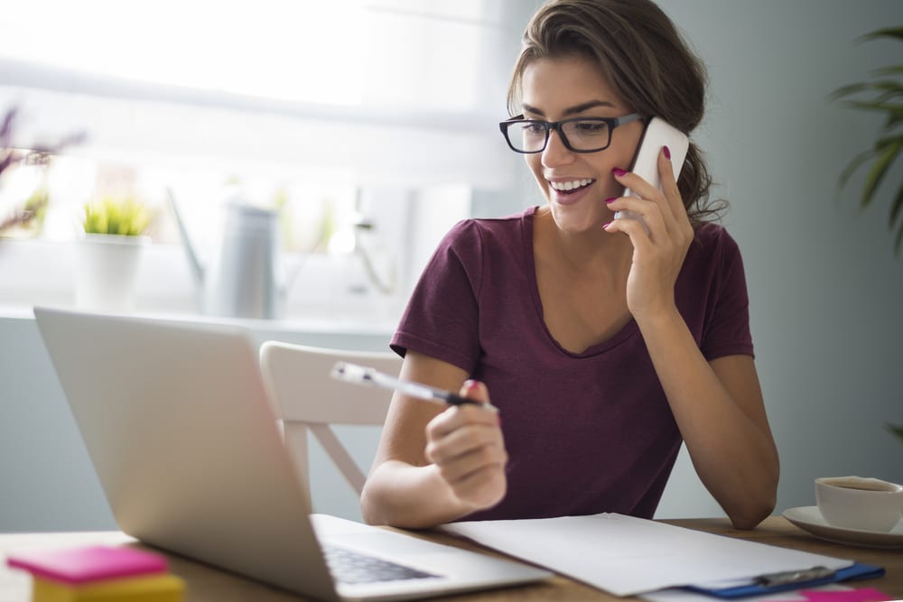 woman wearing glasses talking on the phone and working on a laptop